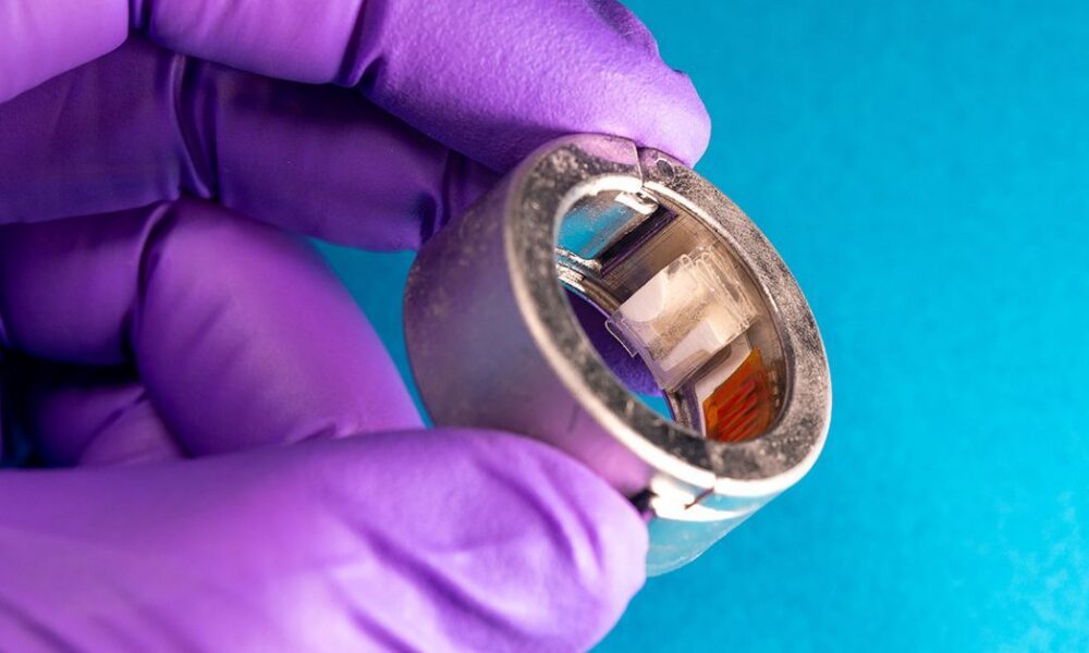 Une bague connectée suit le glucose et les cétones dans la sueur Main gantée de violet tenant un dispositif annulaire métallique avec composants.